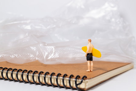 Tiny surfer figure stands on a notebook beside a sheet of plastic, symbolizing environmental issues, pollution, and ocean awareness in a creative concept scene.の写真素材