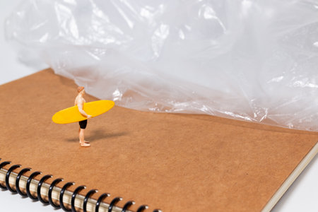 Tiny surfer figure stands on a notebook beside a sheet of plastic, symbolizing environmental issues, pollution, and ocean awareness in a creative concept scene.の写真素材