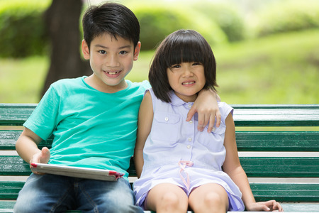 Asian cute boy and little girl are smile and looking the camera, sitting on a wooden bench in the parkの写真素材
