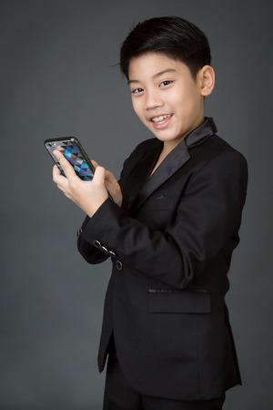 Happy asian child in black suit holding digital cell phone on gray backgroundの写真素材