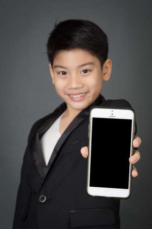Happy asian child in black suit holding digital cell phone on gray background, Focus on screen of phoneの写真素材