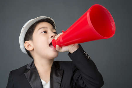 Little asian boy in back suit using megaphone shouting on gray background