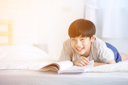 Asian boy rest on the bed and reading book with smile face at home, and sun light in the morning.