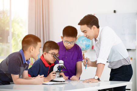 Cheerful male Teacher Explaining Something to a Group of Young Students Inside the Classroom. Asian aboy examining preparation under the microscopeの写真素材