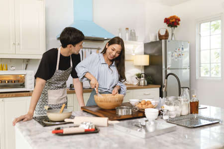 Asian couple Help each other to make a bakery In a romantic atmosphere in the kitchen at home. Young people work together to mix the ingredients in a wooden bowl before stirring them together.の写真素材