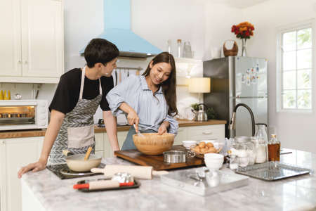 Asian couple Help each other to make a bakery In a romantic atmosphere in the kitchen at home. Young people work together to mix the ingredients in a wooden bowl before stirring them together.の素材 [FY310166927689]