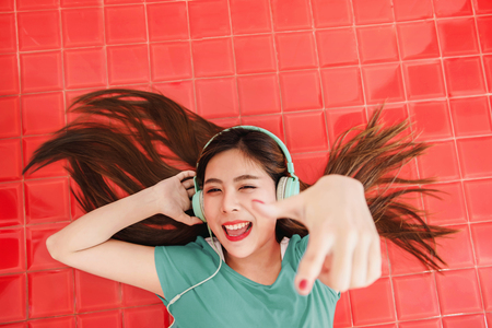 Happy Young Woman Lay on the Red Floor to Listening a Song in Headphone, Looking at camera with Smile. Girl Enjoying Music in House, Top Viewの写真素材