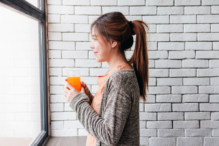Happy Smiling Woman Relaxing in Cozy House and holding a glass of Orange Juice. Side View, Looking Outside windowの写真素材