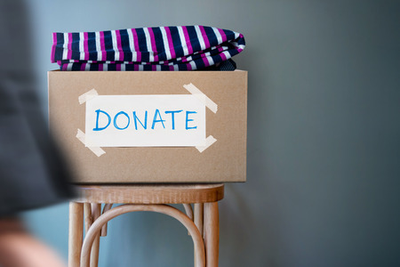 Donation Concept. Donate Box with Used Old Clothes on Chair against Wall in Public Space. blurred walking people as forgroundの写真素材