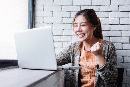 Blurred image of Happy excited Young Woman celebrate in cheerful posture when see a good news in Computer Laptop. Sitting nearby Window in House. Successful and Achievement Conceptの写真素材