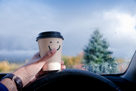 Happy and Positive Mind Concept. Driver holding a Coffee Mug with Smiling Face Cartoon, Blurred Rain in Bad Weather as outside view. Smile on Rainy Dayの写真素材