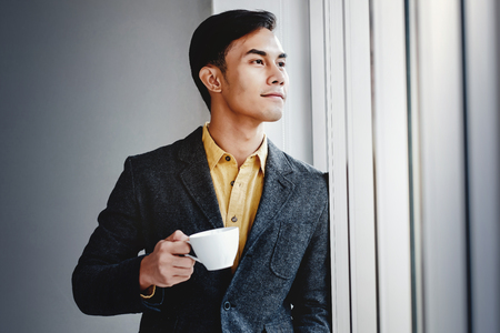 Portrait of Happy Businessman Standing by the Window in Office. Looking Away and Smiling. Dreaming for Success. Drinking Hot Coffeeの写真素材