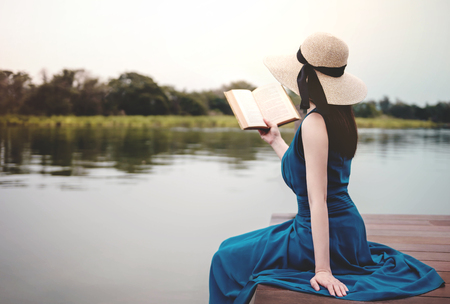 Young Woman Relaxing by Riverside. Sitting on Deck and Reading Book. Unplugged Life Conceptの写真素材