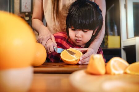 Happy Cute 3-4 Years Old Girl with her Mom Slice some Orange on Wooden Table in Pantry Room. Young Girl is Learning Cook with her Mother. Fruit and Vegetable for Kids Conceptの写真素材