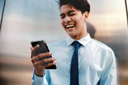 Blurred of a Happy Young Businessman Using Mobile Phone in the Urban City. Lifestyle of Modern People. Front View. Standing by the Wall. Selective Focus on Smartphoneの写真素材