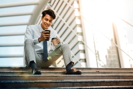 Happy Young Businessman Sitting on Staircase and Using Smartphone. Urban Lifestyle. Low Angle View....