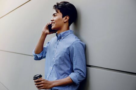 Portrait of a Happy Young Businessman Using Mobile Phone. Lifestyle of Modern People. Standing by the Wall with Coffee Cupの写真素材