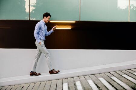 Happy Young Businessman in Casual wear Using Mobile Phone while Walking by the Urban Building Wall....