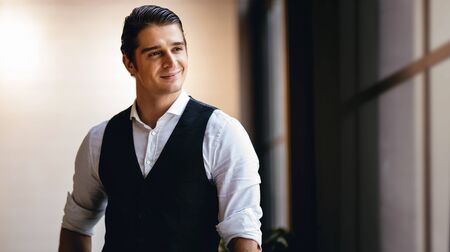 Portrait of a Young Confident CEO or Leader in the Modern Workplace. Smiling Businessman in Comfortable Posture. Standing by the Window and Looking outsideの写真素材
