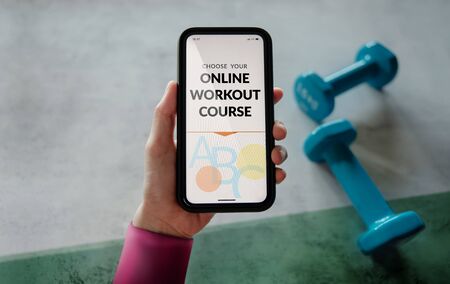 Learning Online Workout Concept. Young Woman using Mobile phone to choosing an Exercising Course. Dumbbell on the floor as backgroundの写真素材