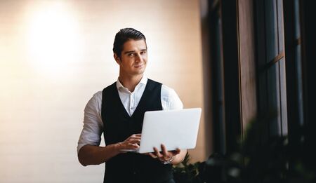 a Young Caucasian Businessman Working on Computer Laptop in the Modern Workplace. Smiling Businessman Standing by the Window and Looking into the Cameraの写真素材