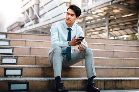 Young Striving Asian Businessman Sitting on Staircase in the City. hand holding a Mobile Phoneの写真素材