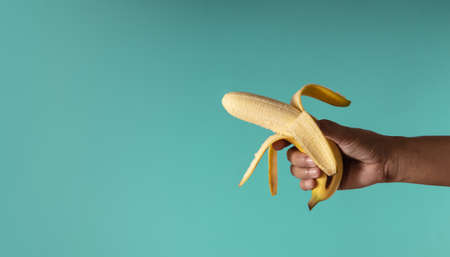 Banana Concept image. Hand Holding a Banana Peel against the blue background, Look like a Gun. Metaphor Photo. Clean and Minimal styleの写真素材