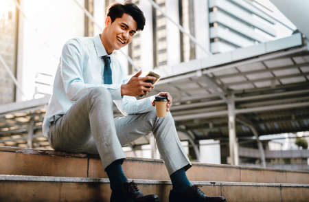 Smiling Young Asian Businessman Using Mobile Phone in the City. Sitting at the Staircaseの写真素材