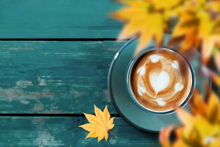 Drinking Coffee in Fall and Autumn Season. Hot Coffee Latte Cup on Blue Wooden Table. Top View. Focus on Latte Art in Heart Shape. blurred Yellow Maple Leaf as foregroundの写真素材