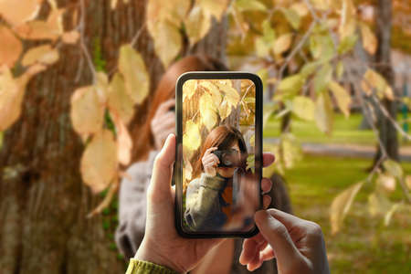 Taking Photo via Smartphone. Young Person Using Mobile Phone to Taking a Photograph for Friend in Outdoor Park.Model on Screen with Digital Camera do the same. Front Viewの写真素材