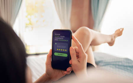 Customer Experiences Concept. Hotel and Resort Review. Happy Client Lay on Bed and Using Smartphone to Giving Five Star Rating for Online Satisfaction Surveys. Positive Feedback on Mobile Phoneの写真素材