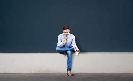 Smiling Young Asian Businessman Using Mobile Phone in the City. Sitting against the Empty Wall