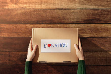 Donation Concept. Woman with Box of Things with Donate label and Heart. Preparing on Wooden Table at Home. Top Viewの写真素材