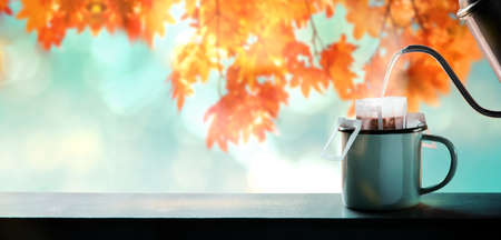Enjoying Coffee in Fall and Autumn Season. Making Hot Coffee by Pouring Water from Kettle into an Instant Coffee Drip Bag. blurred Maple Leaf Tree as backgroundの写真素材