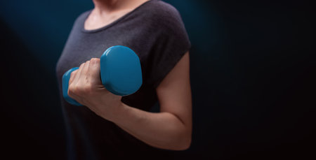 Exercising, Workout with Dumbbell. Sport and Recreation Concept. Close-up of Woman with blue Dumbbell in Dark Room at Home or Gymの写真素材