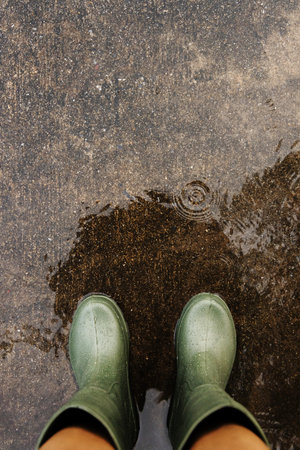 POV shot of person with rain boots standing on wet ground during rainfall, with raindrops making ripples.Rainy season, weather, and environmental awarenessの写真素材