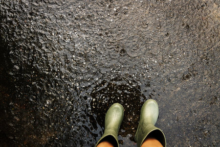 POV shot of person with rain boots standing on wet ground during rainfall, with raindrops making...