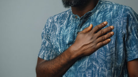 A serious man places his hand on his heart, showcasing an emotional gesture. He is wearing a blue patterned shirt against a neutral gray background.の素材