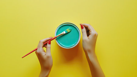 A top view image featuring hands skillfully holding a paintbrush above a bowl of vibrant blue paint against a striking yellow backdrop, capturing the essence of creativity.の素材