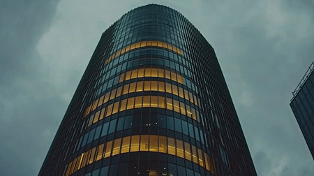 A striking view of a modern skyscraper with a glass facade standing tall against a moody, cloudy sky. The warm lights inside contrast beautifully with the cool exterior, creating a dynamic urban scene.の素材