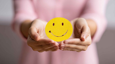 Hands display a cheerful yellow smiley face paper cutout, symbolizing joy and positivity. Ideal for wellness and mental health themes, this image radiates happiness.の素材