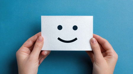 A pair of hands holds a simple card featuring a smiley face, set against a vibrant blue background. This image conveys happiness and positivity, perfect for various applications.の素材