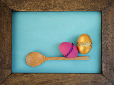 Gold and pink Easter egg and wooden spoon in Wooden frame over blue backgroundの写真素材