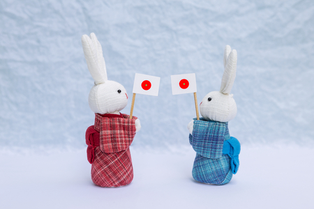 Cute little fabric rabbit doll in red and blue traditional Japanese dress holding Japan flag over blurred white background, decoration object, Japan tourism conceptの写真素材