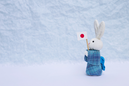 Cute little fabric rabbit doll in blue traditional Japanese dress holding Japan flag over blurred white background, decoration object, Japan tourism conceptの写真素材