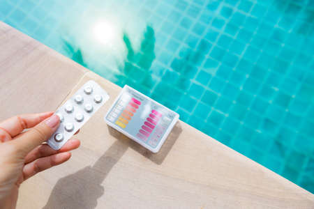 Tablets in girl hand with water testing kit on swimming pool edge with space on blurred clear blue water background, pH and Cl testing, water quality test, pool tester, summer outdoor day lightの写真素材