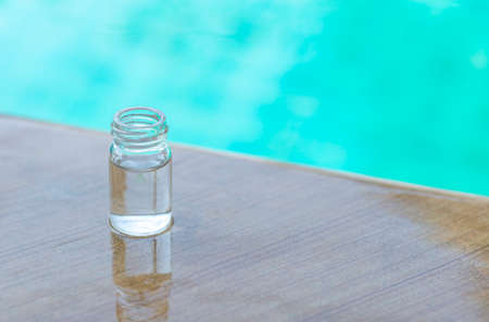 Small glass bottle with clear water on swimming pool edge over blurred blue water background, water...