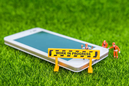 Work in progress sign with miniature worker and smartphone on green grass background, technology...