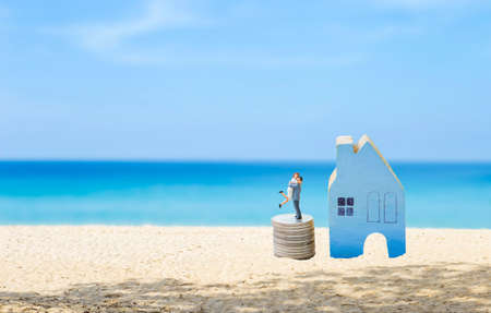 Miniature couple on stack of coin with blue wooden hosue model on the beach with space on blue sea backgroundの写真素材