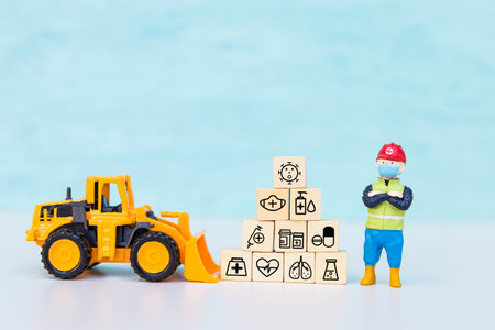 Miniature worker wearing safety helpmets with medical icon cube and yellow front loader truck with...